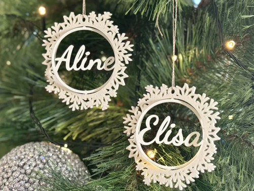 Duas bolas de Natal em tom claro com contorno recortado de flocos de neve e os nomes Aline e Elisa, penduradas numa árvore iluminada.