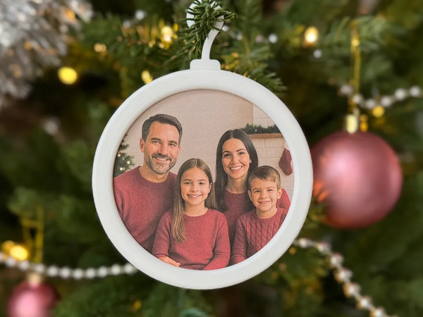 Bola de Natal circular com moldura branca e fotografia de quatro pessoas, pendurada numa árvore de Natal iluminada.